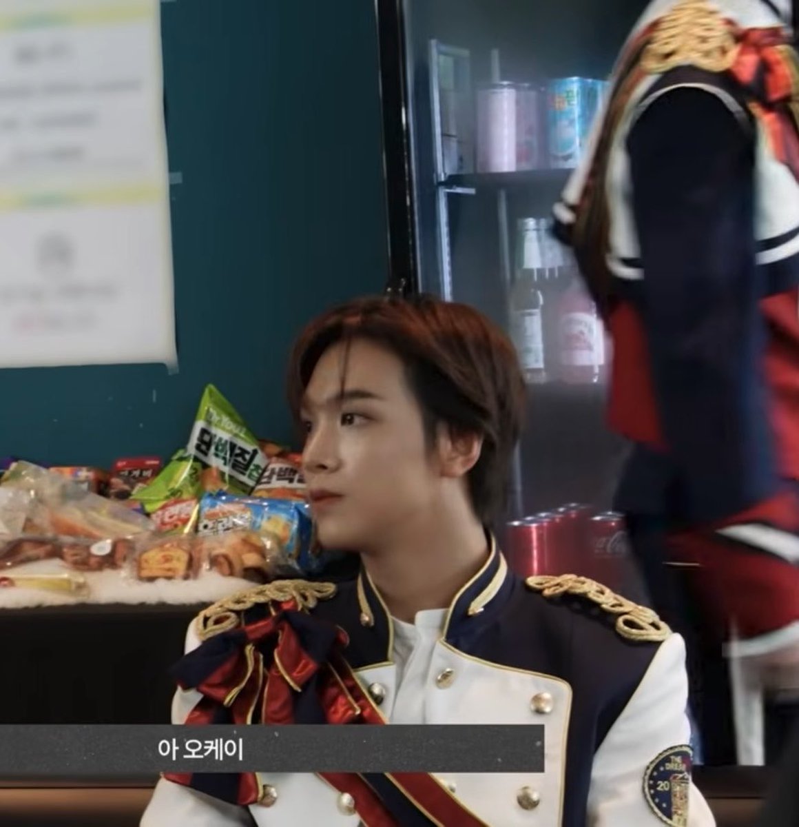 A young man in a regal navy and white uniform with gold epaulettes and a red bow, surrounded by snacks and Coca-Cola cans, with the Korean caption '아오케이'.