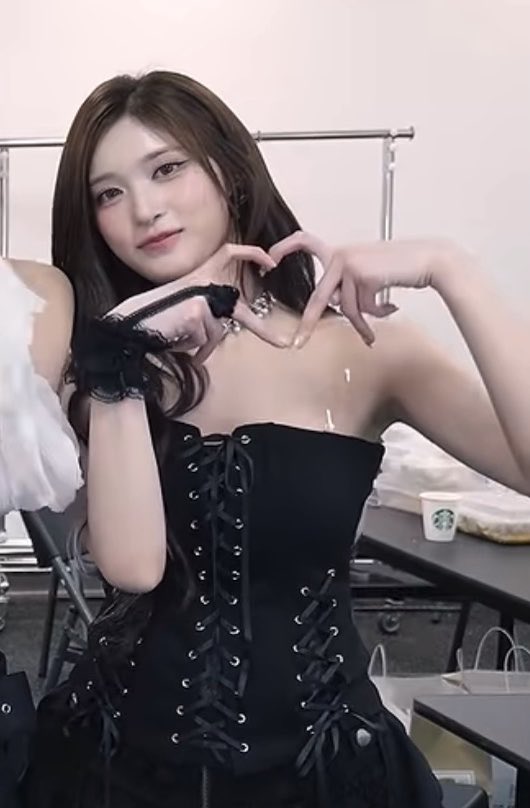 A young woman with long brown hair poses backstage, making a heart gesture with her hands while wearing a black lace-up corset dress.