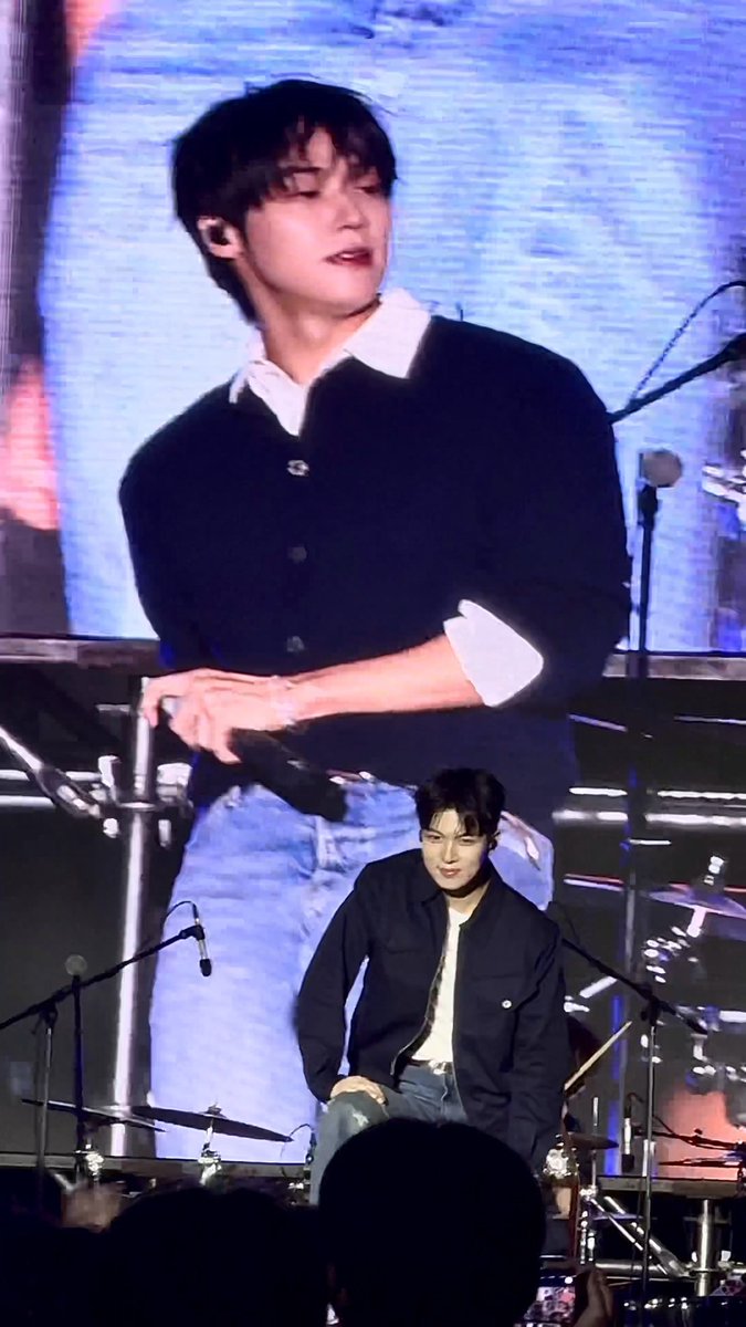 A man in a black cardigan and blue jeans sits on a stage in front of a drum set, with a large screen behind him showing a close-up of his image.