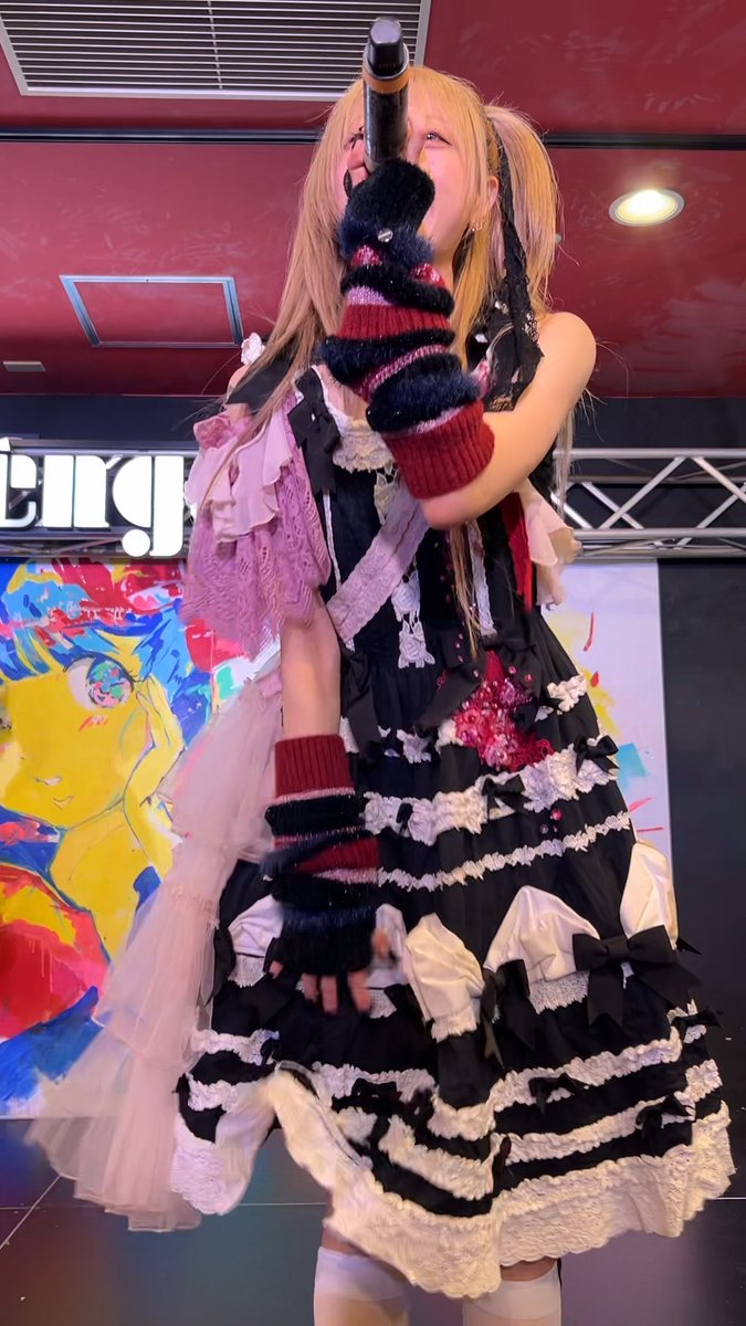 A young woman cosplaying in a gothic lolita black and pink dress with striped fingerless gloves, holding a prop gun while posing in front of a colorful poster.