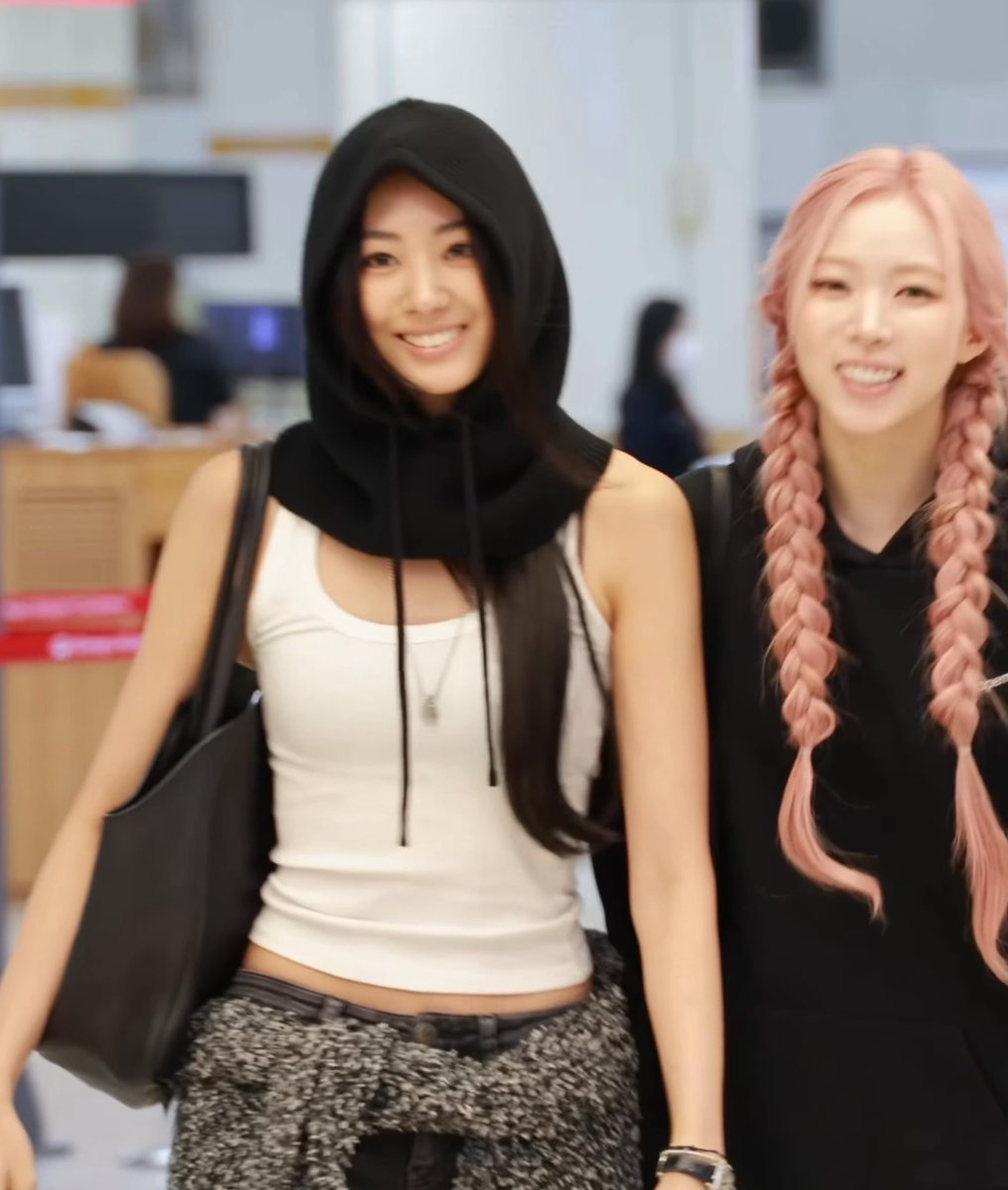 Two young women smile at the camera; one wears a black hood over a white tank top and carries a large black bag, while the other has pink hair styled in twin braids and wears a black hoodie.