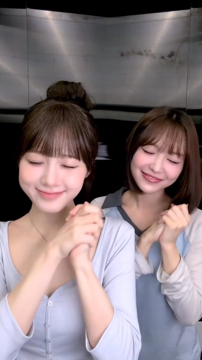 Two young women with similar hairstyles pose with closed eyes and smiles, holding each other's hands in front of a dark background.