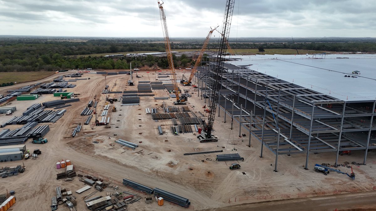 JoeTegtmeyer's tweet photo. Giga Texas Optimus factory construction update with 2 short video clips, one 360 at medium-altitude & a closer, lower flight showing tight views of the main steel structure!

Ground work started in late March, with 1st steel on May 27. By 1 October,  the frame was nearly at the north grade beam, large sections of upper floors were poured, & roofing insulation is arriving. The E side & the entire S half of the foundation have footings & grade beams, but no steel yet. The basic steel structure is ~ 40% assembled so far.

This is the dedicated high-volume 7+ million sq ft (~ 650,000 sq m) Gen 4 Optimus plant. @Tesla has not given a standalone cost, but 2026 company capex was guided at ~ $20–25 billion for AI, robotics, & factory expansion; outside estimates put the broader N Campus at ~ $5–10 billion.

This is in addition to the Fremont California Optimus plant (which is the Gen 3 pilot line) is sized at ~ 1 million units a year, expected to begin production in late 2026.

The Giga Texas plant is 10X larger & has a planned capacity of ~10 million robots a year at full scale (a long-term ceiling, not the startup rate). The current target for limited initial production is late 2027.

What is still ahead for construction over the next 6 to 8 months? Electrical conduit, water & sewage pipes, fire-water loop, storm water management drainage & underground pipes, exterior walls, roofing, ground slab & upper floor concrete & mechanical rough-in then equipment deliveries & installation. As you can see, lots of progress already, but there is a lot left to do before production line setup can happen.