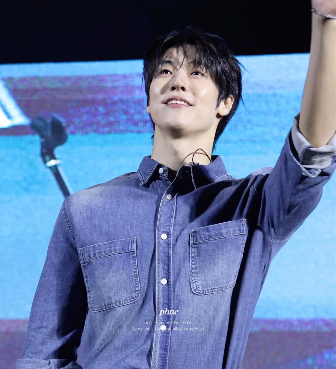 A young man with dark hair smiles brightly on stage, raising one arm while wearing a denim shirt, with the watermark "phme for N.FLYING SEUNGHYUB" visible. The image captures Lee Seunghyub of N.Flying performing at the JAU:ON festival at Jangan University on October 1, 2026, matching the post's caption about his glowing face.