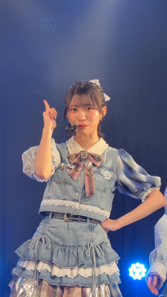 A young woman performs on stage in a denim outfit with a white lace collar, plaid bow, and ruffled skirt, pointing her finger upward.