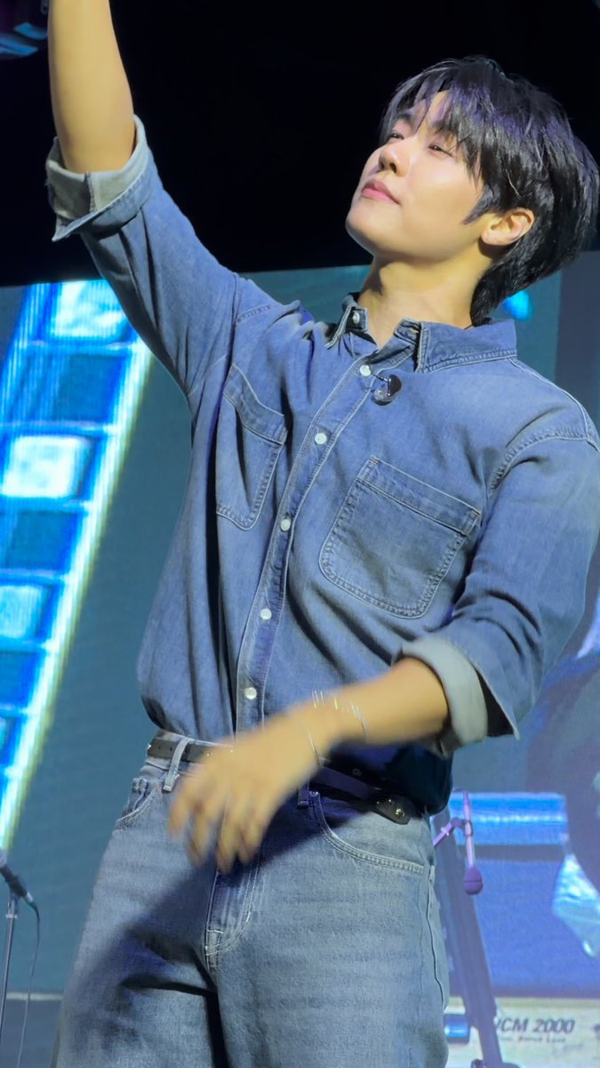 A young man with dark hair, wearing a denim shirt and jeans with a lapel microphone, performs on stage with his arm raised dramatically. In the bottom right corner, partially visible text reads "MC 2000," and the caption captures his endearing, animated energy.