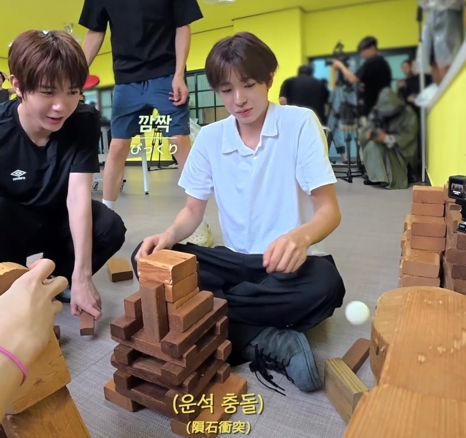 A young man in a light blue polo shirt sits cross-legged on the floor, intensely focused as he carefully removes a wooden block from a towering Jenga-like stack, while another person in black crouches beside him watching closely. Korean text overlays read "까칙 님 잘 다녀 오세요" and "(운석 충돌)(隕石衝突)" at the bottom, and the tense concentration on the player's face perfectly captures the nervous, trembling anticipation described in the caption—stunned yet silent like a buzzing dog.