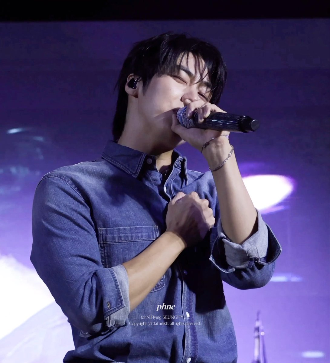 The post appears to be from a fan account sharing a photo of Seunghyub from the K-pop band N.Flying performing at a university festival. The image shows the vocalist passionately singing into a microphone on stage, wearing a denim shirt and in-ear monitor, with his hand pressed to his chest — visually capturing the heartfelt lyric "I will give you my heart completely" referenced in the caption.