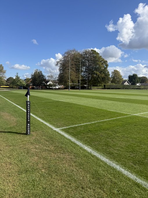A view of a green sports field with a black corner flag reading "DANIELS RHONDDA", with H-shaped rugby goalposts visible in the distance under a blue sky.