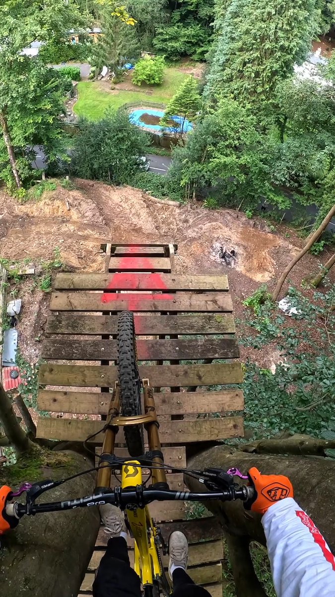 A POV shot from a GoPro HERO13 Black Ultra Wide camera captures a mountain biker perched atop a towering wooden ramp, with the front wheel of their Scott bike dangling over the edge above a backyard pool far below. The rider's orange-gloved hands grip the handlebars as they prepare to drop into what appears to be an extreme downhill stunt, showcasing the dramatic perspective made possible by the ultra-wide lens.