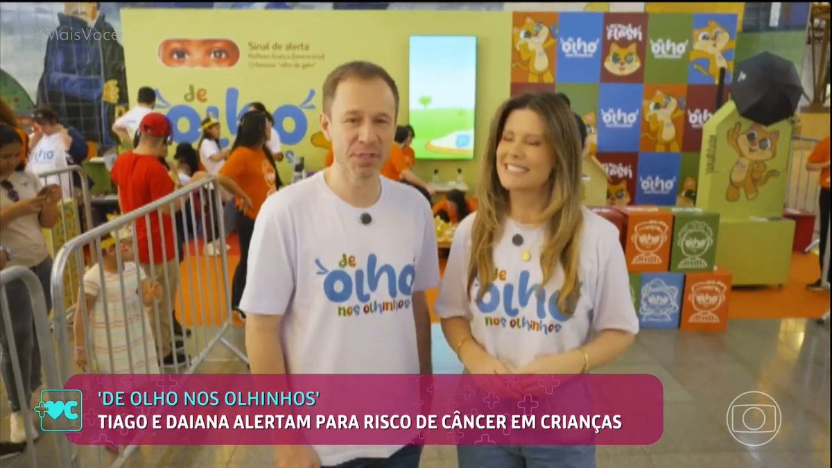 Two news presenters, Tiago and Daiana, stand in front of a children's cancer campaign booth "De Olho Nos Olhinhos" at "Mais Você," wearing matching white t-shirts, with on-screen text alerting parents to the risk of childhood cancer.