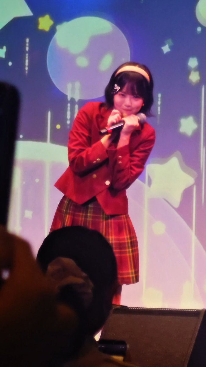 A young woman in a red blazer and plaid skirt performs on stage holding a microphone, with a colorful starry backdrop behind her.