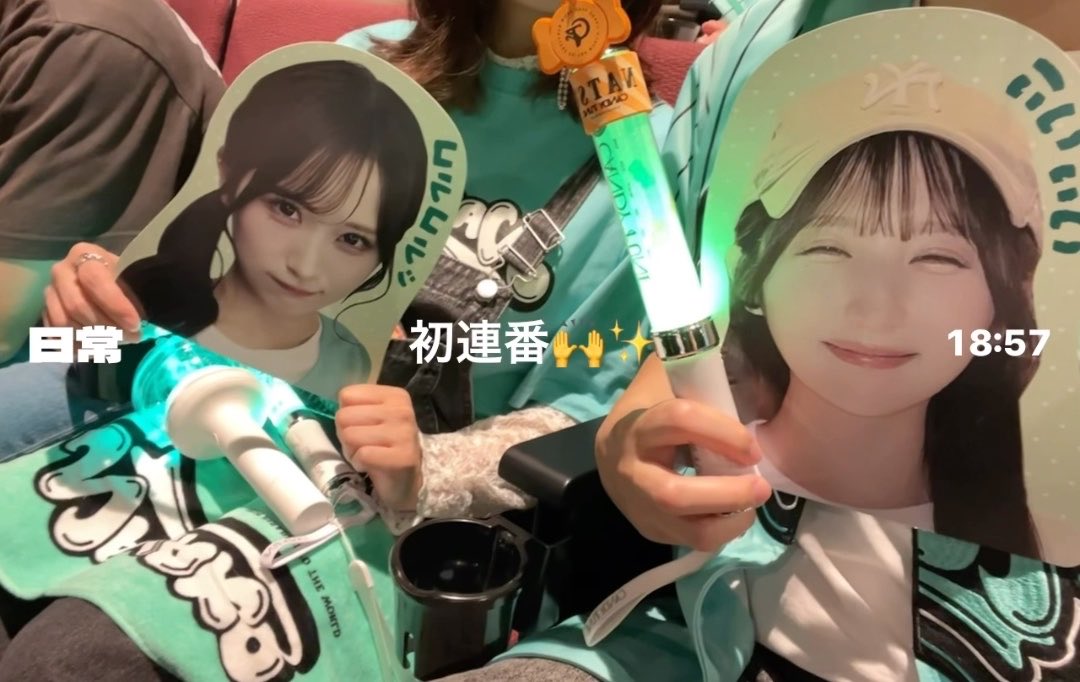 A fan holding illuminated glow sticks (penlights) and cardboard cutout face boards with printed images of idols, with overlay text '日常', '初連番', and '18:57'.