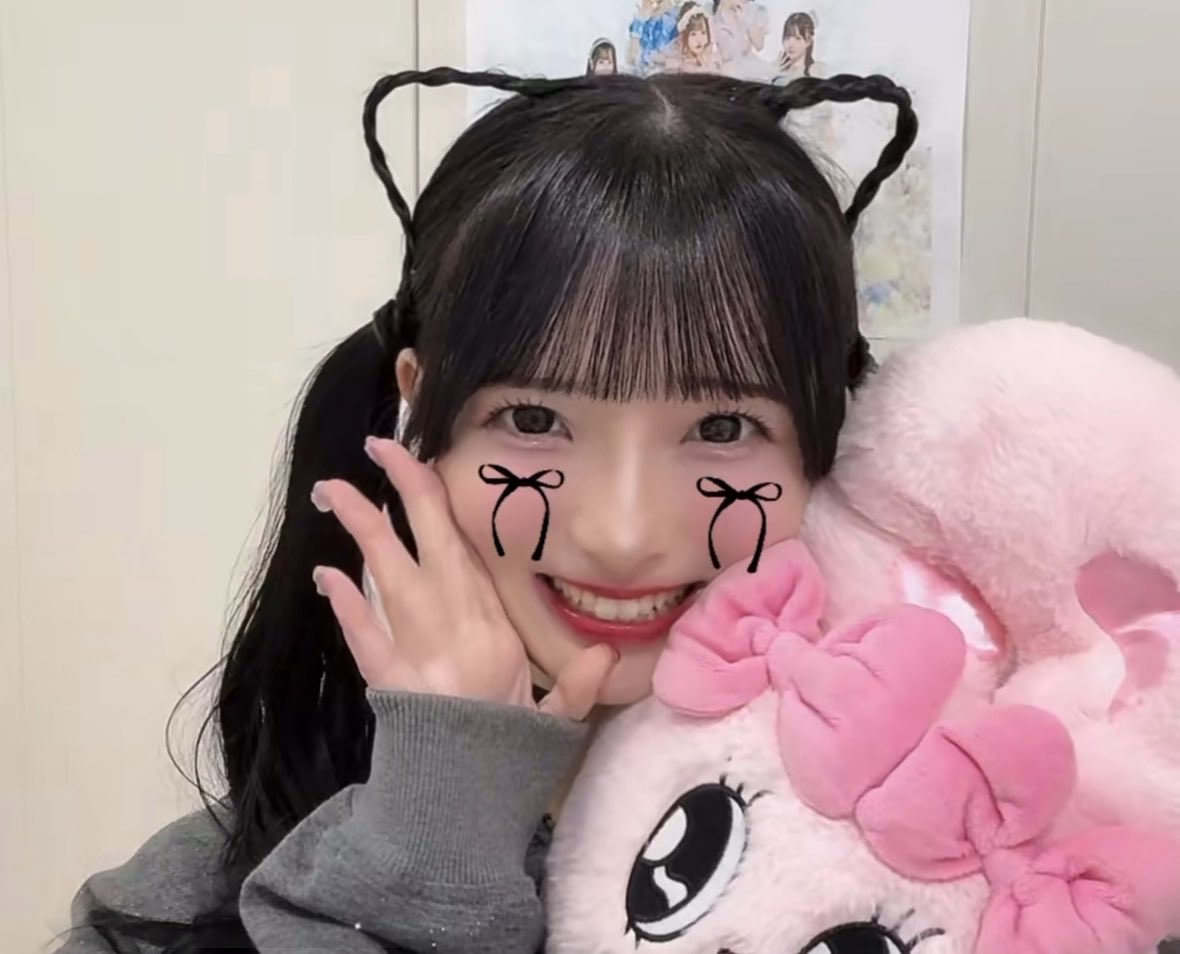 A smiling young woman with dark hair styled in twin braids on top of her head and long pigtails wears digital bow stickers on her cheeks while holding a pink plush toy.
