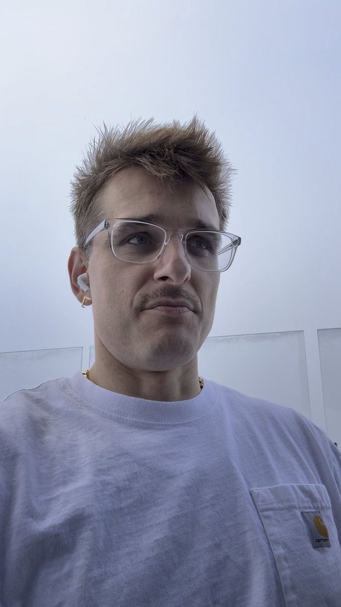 A selfie-style photo shows a man with spiky blonde-brown hair, glasses, and an earpiece, wearing a light purple Carhartt t-shirt against a pale sky background.