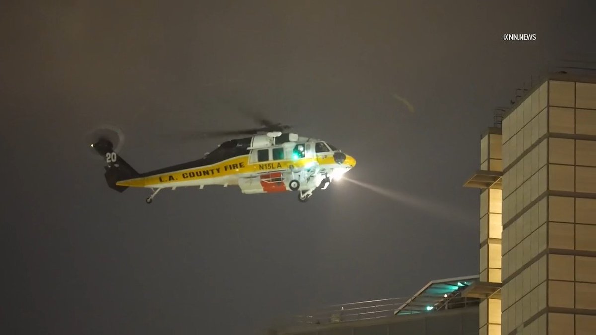 A Los Angeles County Fire helicopter marked "L.A. COUNTY FIRE" with tail number "20" and "N15LA" hovers at night, its spotlight cutting through the darkness. The image likely shows the rescue helicopter responding to the fatal medevac crash off Catalina Island.