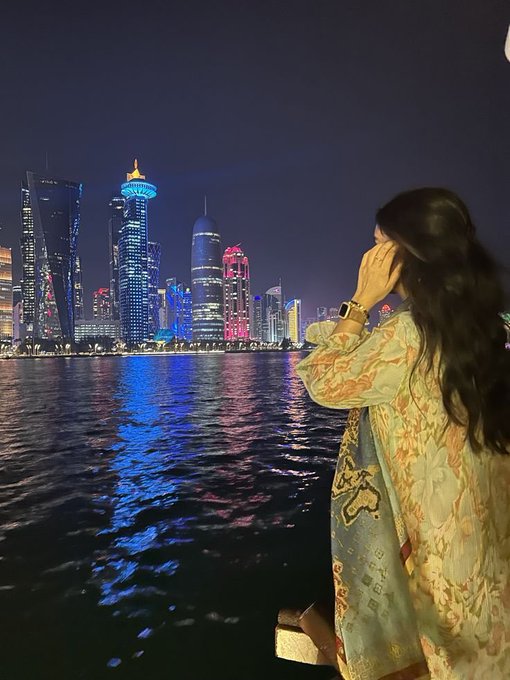 A woman with long dark hair wearing a patterned sheer dress stands by the water at night, looking out at the illuminated Doha, Qatar skyline.