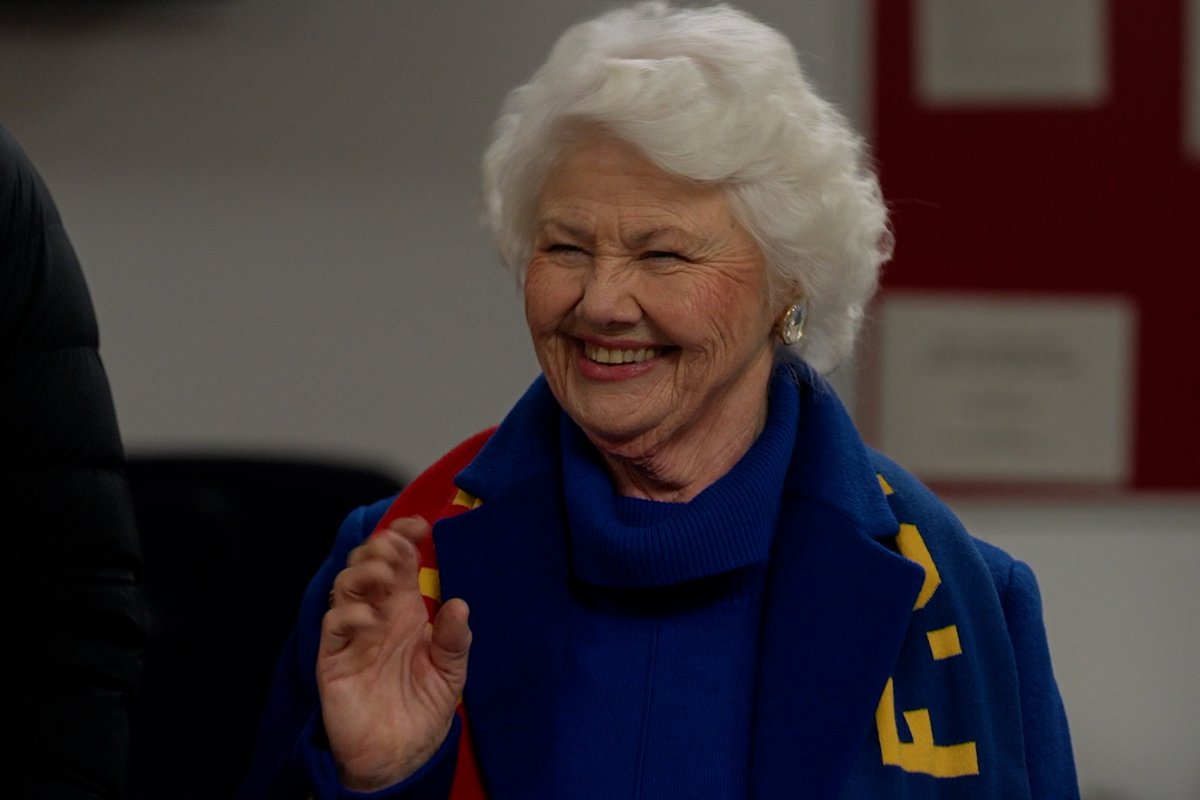 The image shows an elderly woman with white hair, smiling warmly while wearing a blue coat with yellow lettering and a blue turtleneck. This appears to be a still from the TV series "Ted Lasso," likely featuring a beloved character whose meaningful words tie into the post's caption about a new episode now streaming.