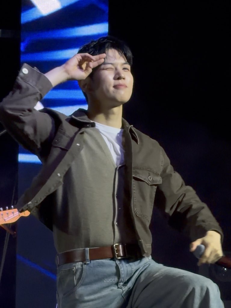 A young man with dark hair wearing a brown jacket and jeans, posing on a stage with one hand near his head, and a watermark 'zucchin_r' is visible at the bottom.