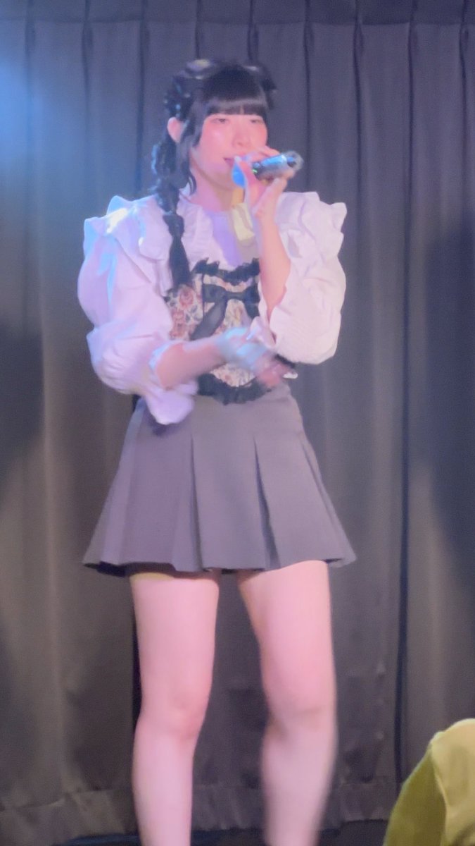A female performer on stage singing into a microphone, wearing a white puffy-sleeved blouse, a floral bodice, a grey pleated skirt, and dark hair accessories.