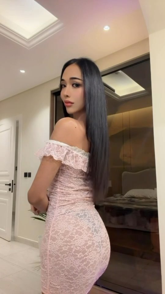 A woman with long dark hair poses in an off-shoulder pink lace dress inside a modern room with a bed visible behind her.