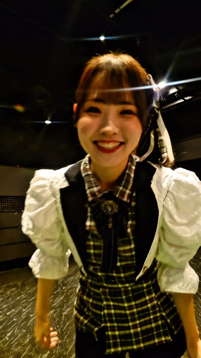 Smiling young woman with shoulder-length brown hair wearing a white puffy-sleeved top with a plaid dress and decorative bow, photographed at night on a city street.