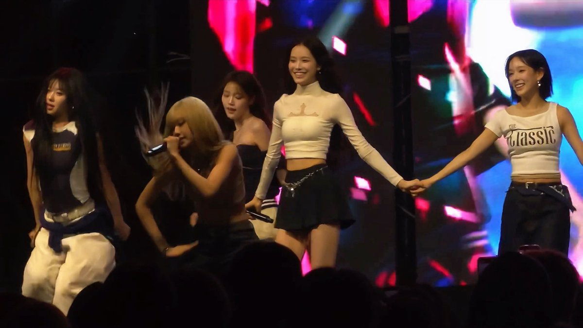 A group of female performers is on stage during a concert, with one member holding a microphone and singing while others dance; one wears a white 'Classic' tank top, and the stage features bright pink and blue lighting.