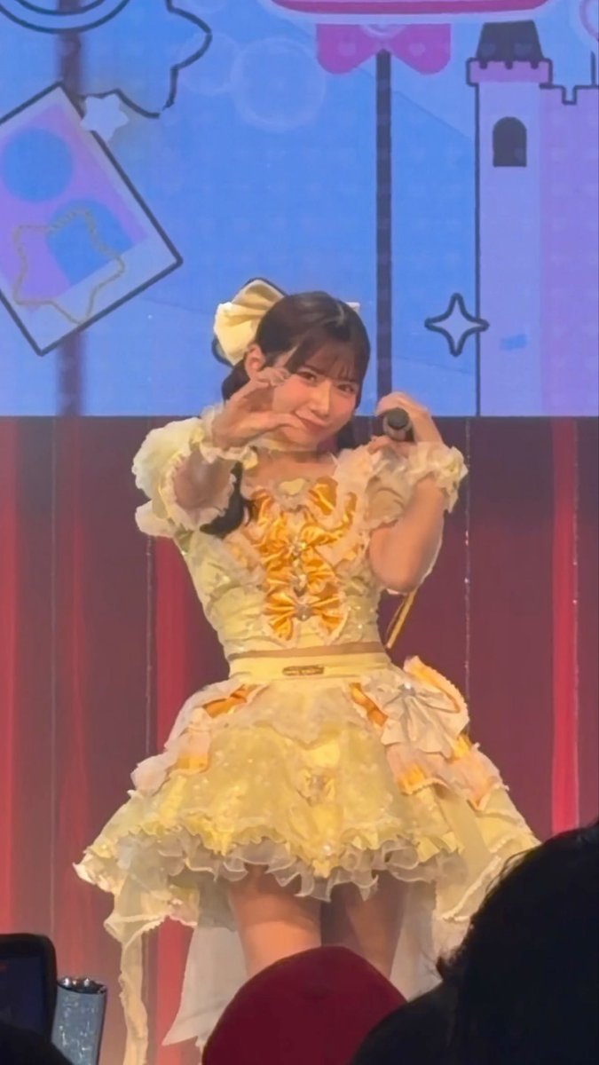 A young female performer in an ornate yellow dress with bows makes a hand gesture while holding a microphone on stage.
