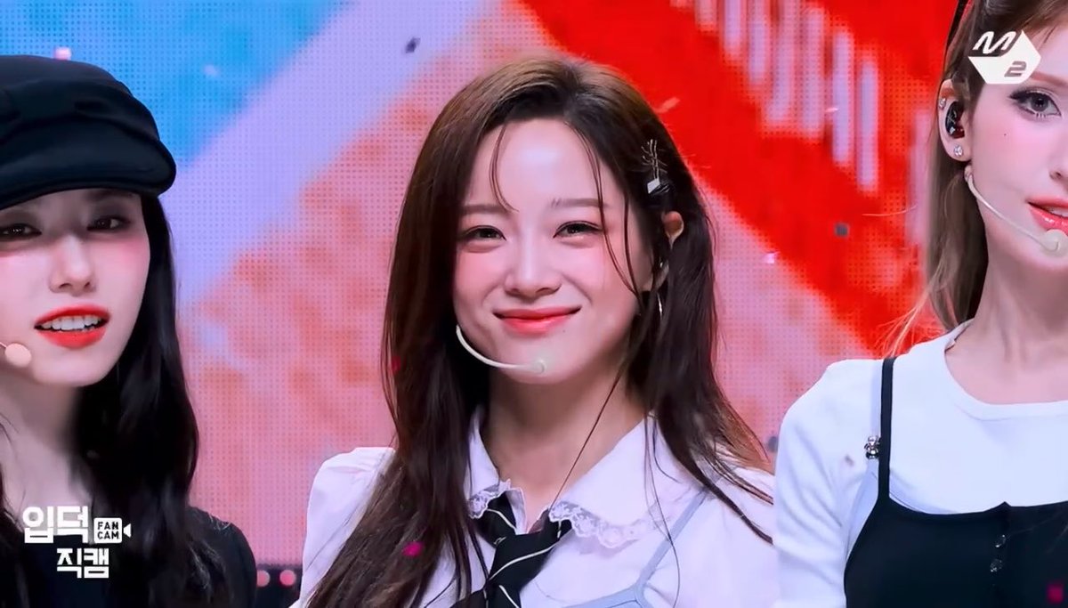 The image shows a smiling young woman with long brown hair wearing a white blouse with a black ribbon, appearing on the South Korean music show "엠카" (M Countdown), with the "M2" logo visible in the corner. Her warm, gentle smile and elegant styling convey the enduring charm and affection referenced in the caption "suddenly is still so loved."