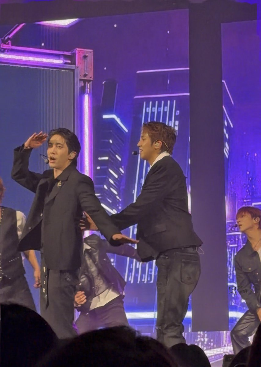 Two male K-pop performers in black suits dancing and singing on a stage with purple lighting and a cityscape backdrop.