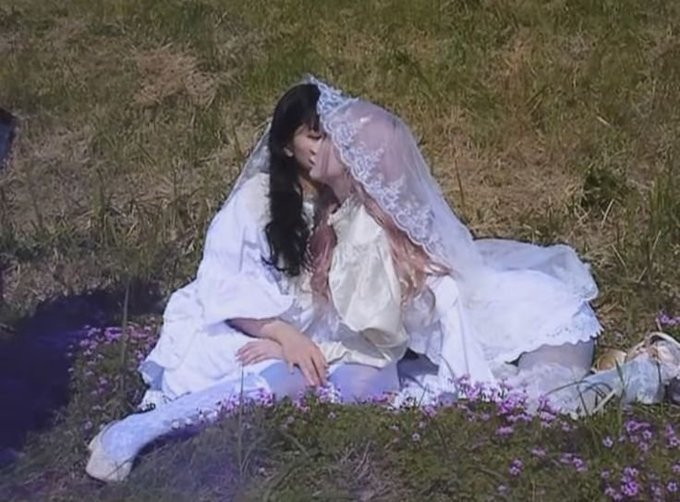 A figure in an elaborate white wedding dress and veil sits on grass surrounded by small purple flowers, with dark hair partially obscuring their face.
