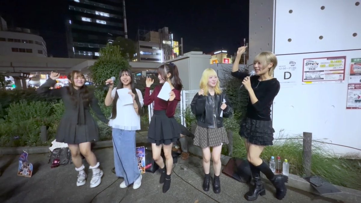 A group of five young women pose and wave during an outdoor nighttime event, with some holding papers and a microphone, in front of a wall displaying a 'D' sign and an open-late notice.