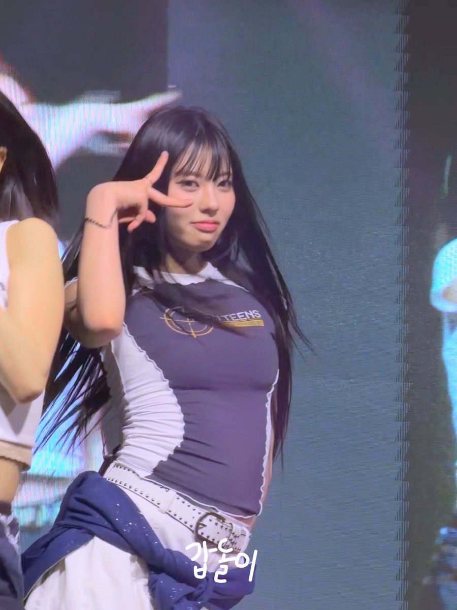 A female performer with long black hair poses on stage, flashing a peace sign and winking. She wears a dark blue and white fitted top with 'TEENS' visible on the chest.