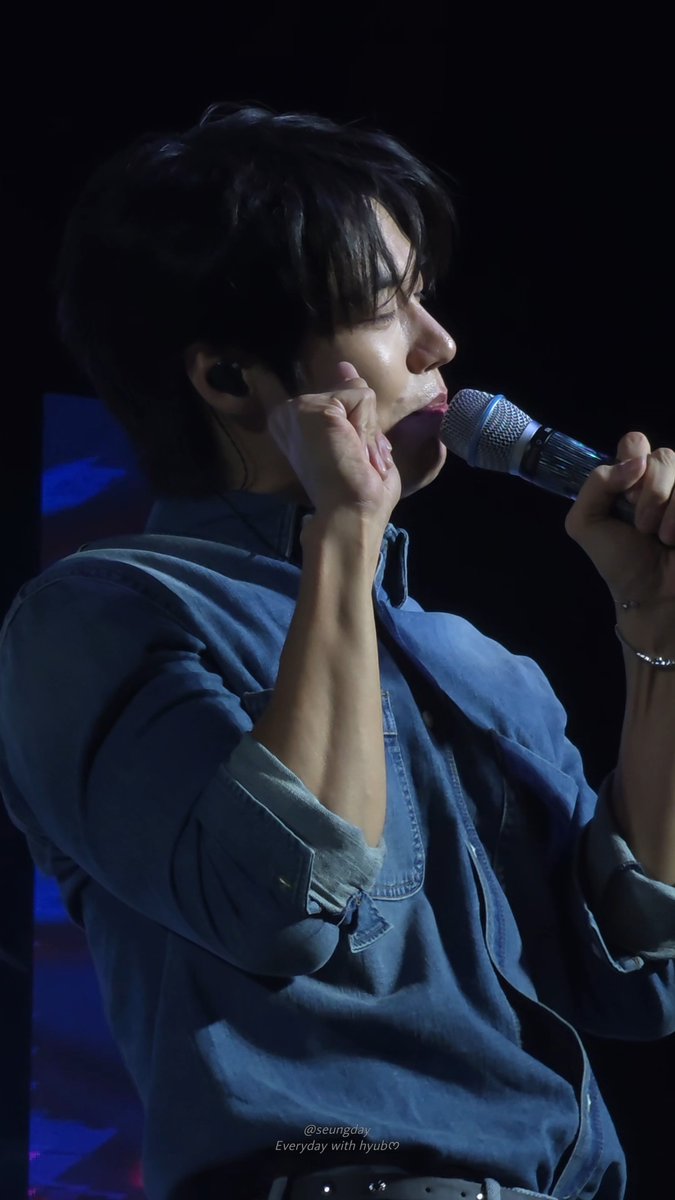 A performer with dark hair, wearing a blue denim shirt and in-ear monitors, sings into a microphone on stage with the caption '@seungday Everyday with hyub♡'.