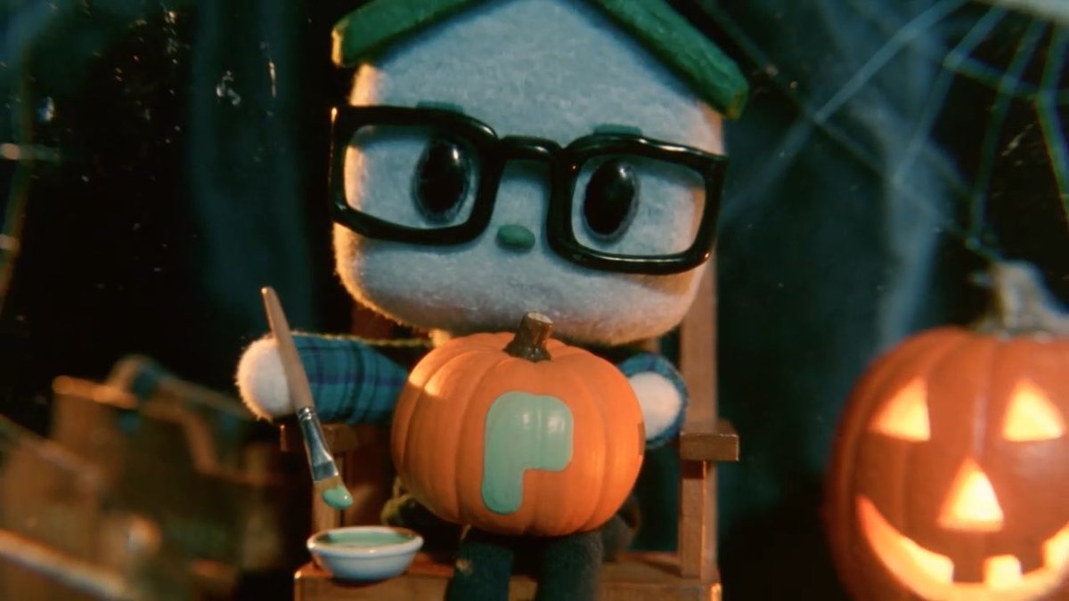 A small plush character wearing black-rimmed glasses and a green beanie sits in a chair, holding a paintbrush and posing with a pumpkin painted with a light blue 'P', alongside a lit jack-o'-lantern in a spooky setting.