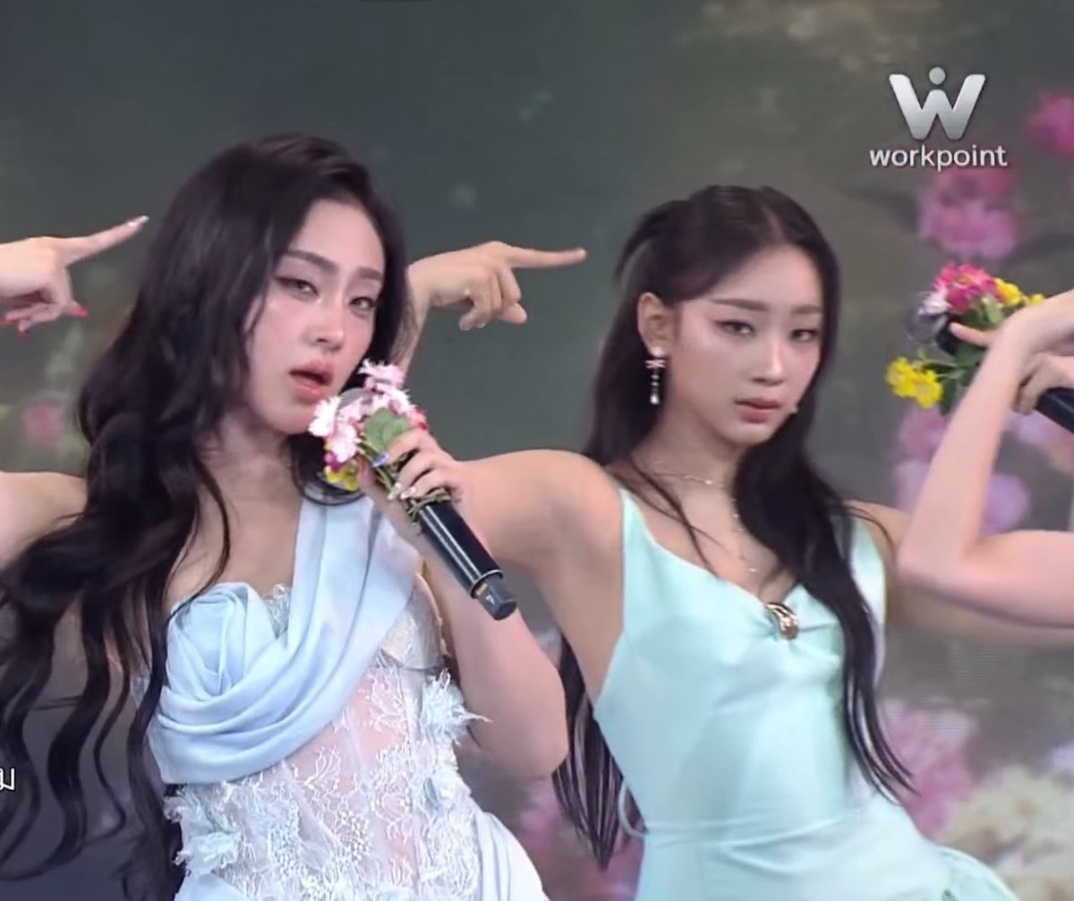 Two female K-pop performers perform on stage, one in a white dress and the other in a mint green top, both holding flower-adorned microphones, with the Workpoint channel logo in the corner.