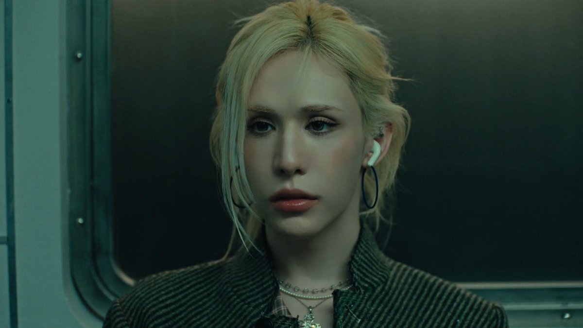 A cinematic close-up of a blonde woman with blue eyes, wearing large hoop earrings, a layered necklace, and an earbud, standing against a dark, greenish background.