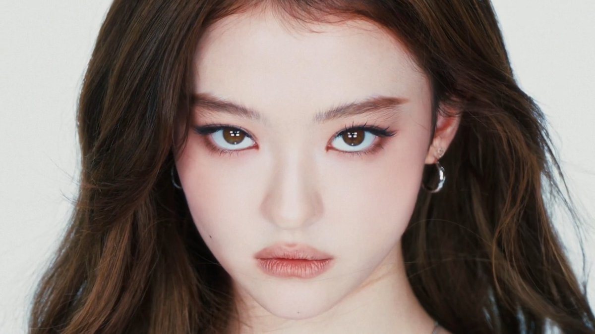 A close-up portrait of a woman featuring detailed eye makeup and hoop earrings against a white backdrop.