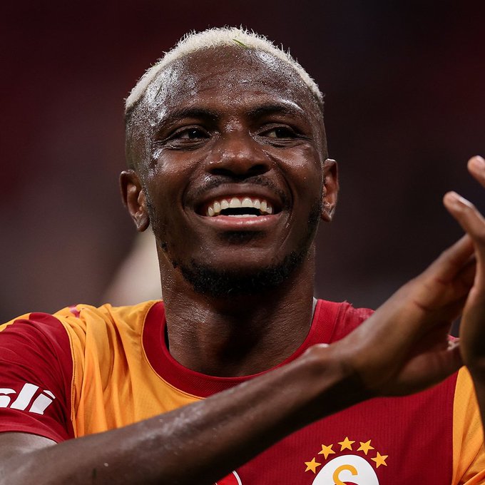 Victor Osimhen, wearing a red and orange Galatasaray jersey with the club's crest and four stars visible, smiles broadly while clapping his hands. The image accompanies news of the Nigerian striker's decreased Transfermarkt market value from €75 million to €65 million, reflecting a shift in his valuation likely tied to his high-profile loan move to the Turkish club.