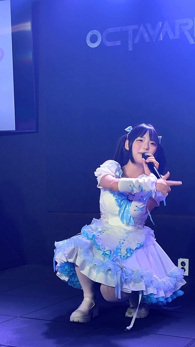 A young performer crouches on a stage bathed in blue lighting, holding a microphone while wearing an elaborate white and blue ruffled outfit, with 'OCTAVAR' visible on the screen behind her.