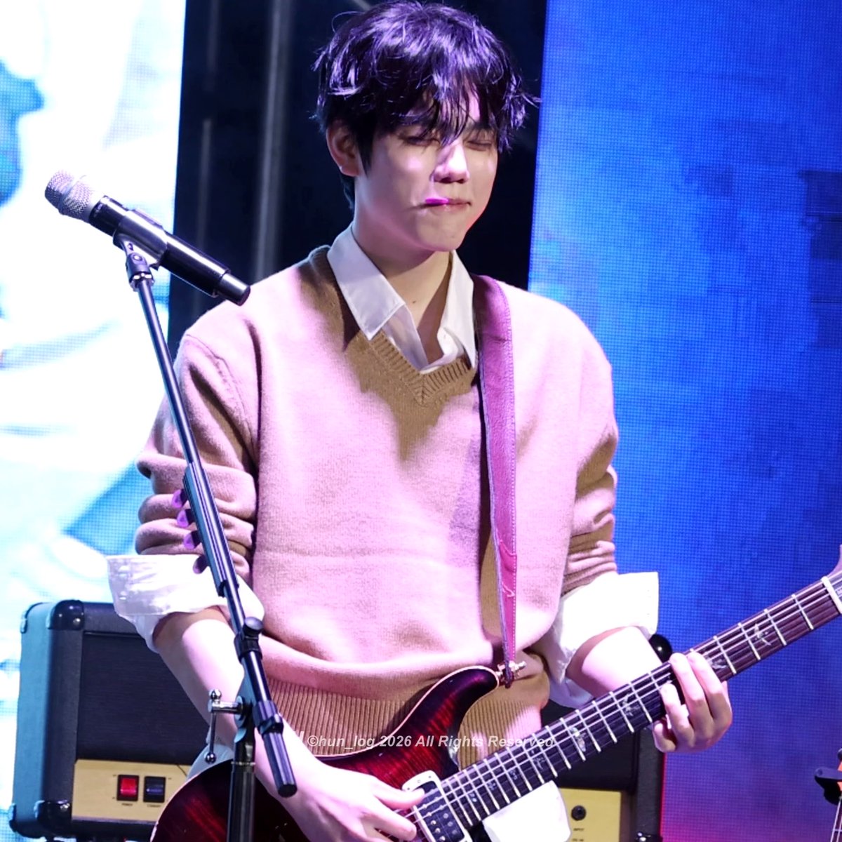 A young man with dark hair plays an electric guitar on stage, wearing a pink sweater vest over a white collared shirt, with a microphone stand in the foreground and a watermark reading "©hun_log 2026 All Rights Reserved" at the bottom.