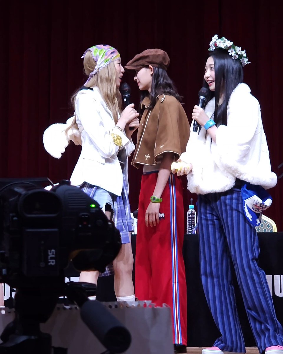 Three young women in colorful outfits, including headscarves, berets, and floral crowns, stand on a stage holding microphones next to a professional video camera.