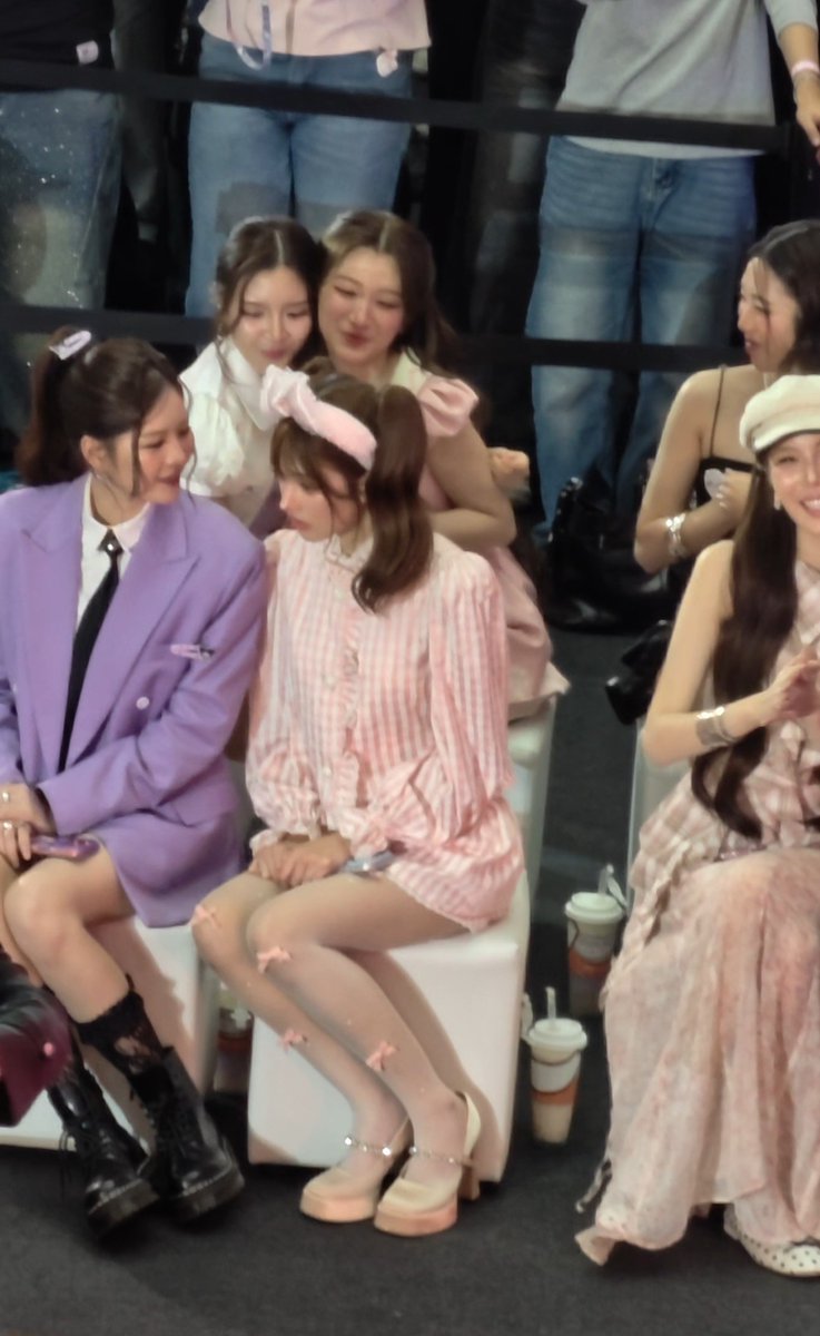 A group of young women in pastel and pink outfits, including one in a purple blazer and others in pink dresses with bows, sitting together on white chairs at what appears to be a fan event.