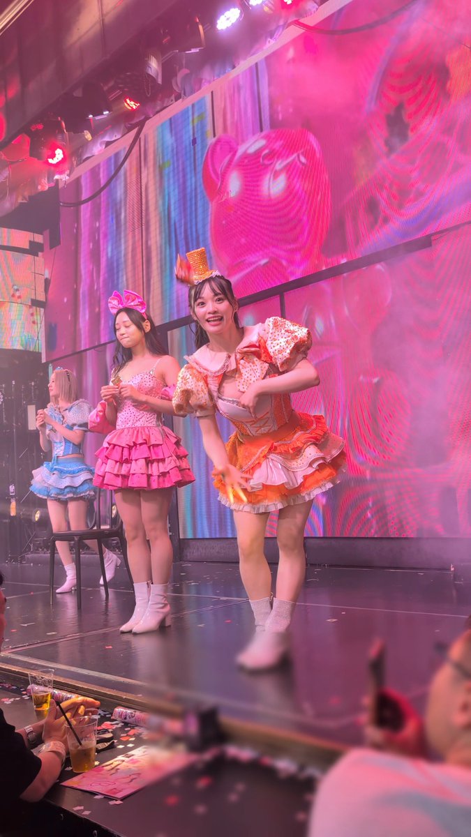 Three female performers in colorful frilly outfits performing on stage at what appears to be an idol cafe, with one in an orange dress bowing, confetti scattered on the floor, and audience members in the foreground.