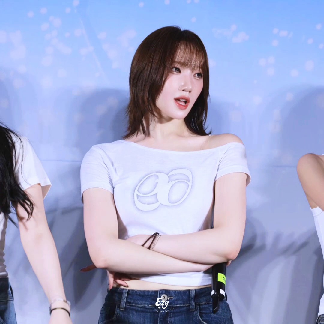 A young woman with shoulder-length brown hair and bangs stands on a stage wearing a white off-the-shoulder crop top with a logo and blue jeans, her arms crossed.