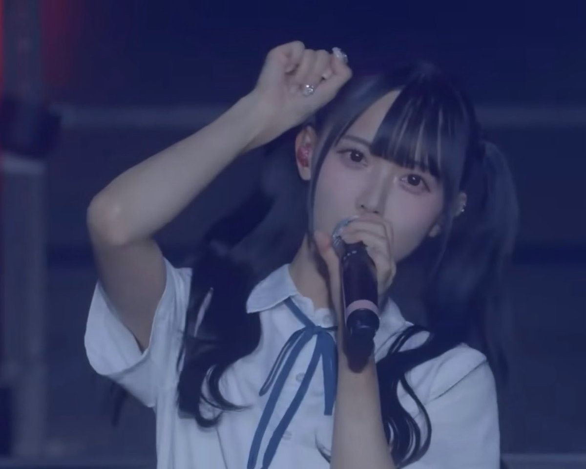 Concert photo of a young female performer with dark pigtails in a white shirt and blue ribbon, singing into a microphone.