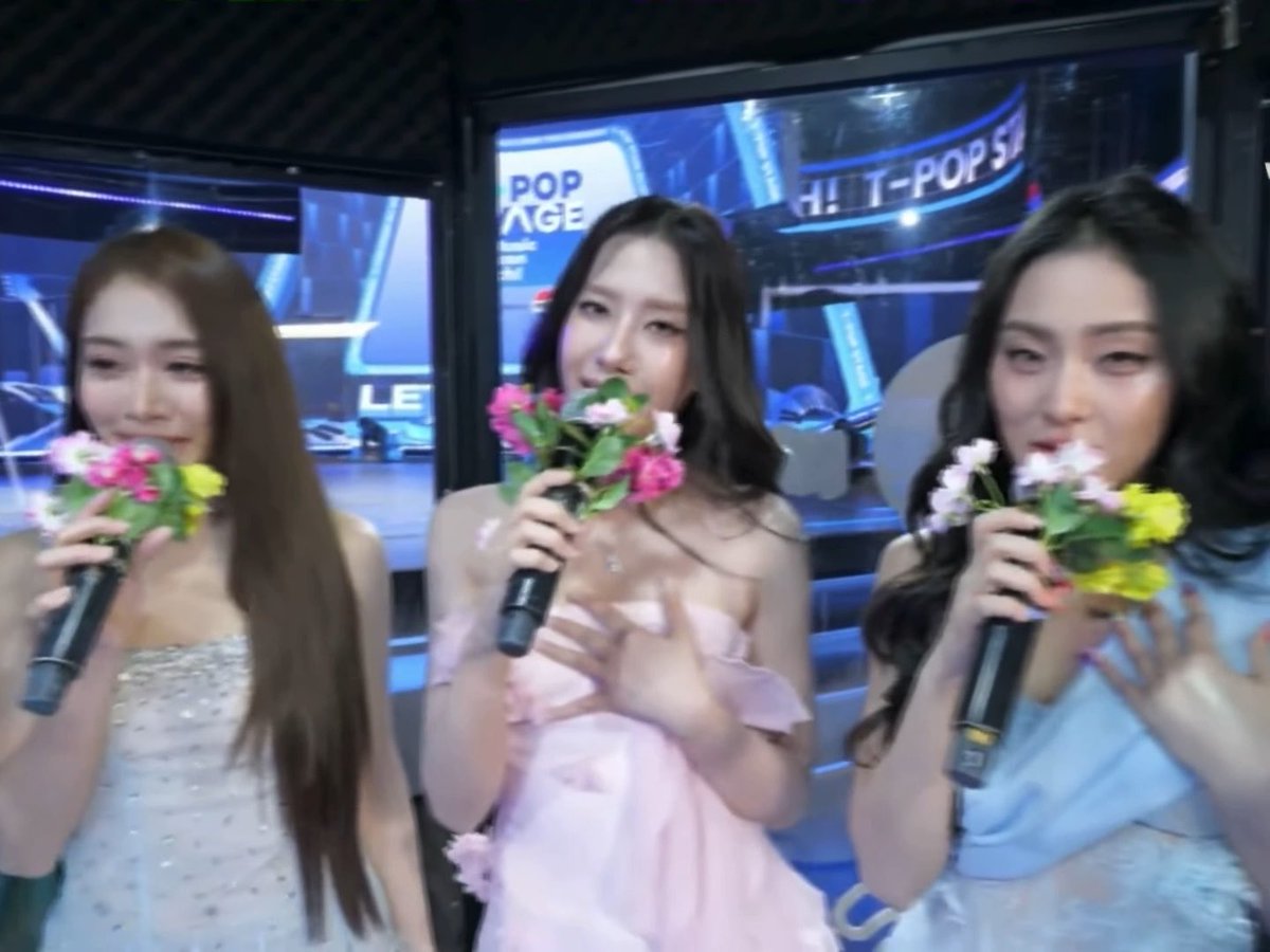 Three women hold flower bouquets and microphones, smiling on a stage with screens displaying "POP STAGE" and "HIT-POP STAGE" in the background.