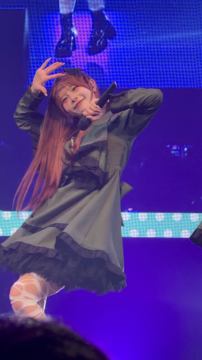 A young female performer with long reddish hair in a black dress poses energetically while holding a microphone on a stage with blue lighting.