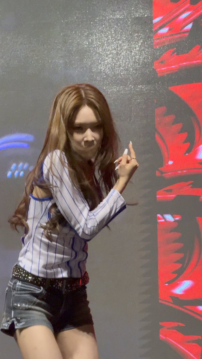 A performer with long auburn hair wearing a white and blue striped jersey and denim shorts, posing on stage with red graphics in the background.