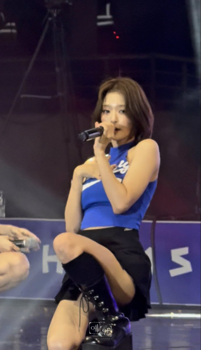 A performer with short brown hair singing into a microphone on stage, wearing a blue sleeveless top, black skirt, and knee-high lace-up boots.